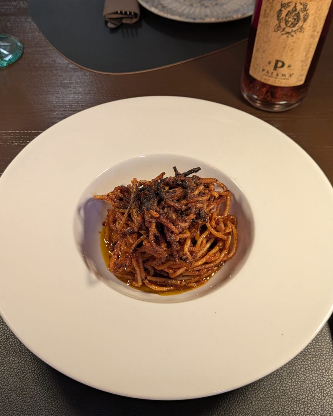 Spaghetti all&rsquo;assassina di Belgusto: pasta rossa e caramellata con la crosta scura del fondo, al centro di un piatto fondo bianco, accanto a una bottiglia di rosso pugliese