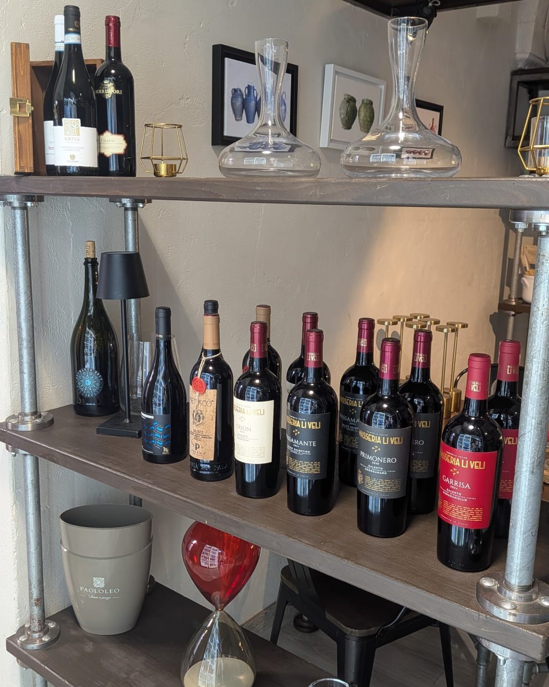Lo scaffale dei vini di Belgusto: due file di rossi del Salento e della Valle d&rsquo;Itria, due decanter di cristallo sul ripiano alto e un magnum accanto a una lampada