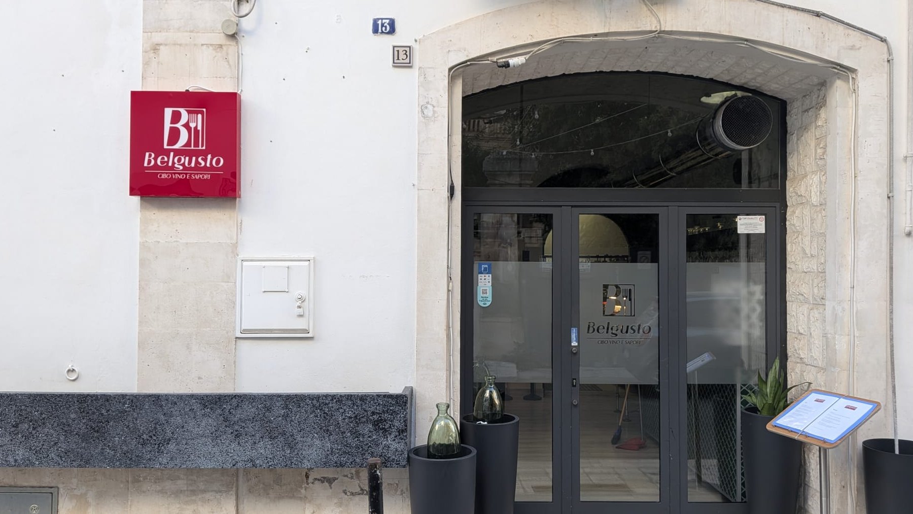 La facciata di Belgusto: l'insegna rossa col marchio sul muro bianco e, accanto, il portone ad arco con le vetrate scure e il marchio inciso