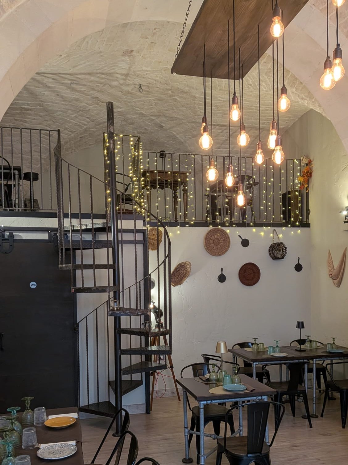 La sala di Belgusto: volta a stella imbiancata a calce, lampadine a filamento appese a una trave, la scala a chiocciola in ferro che sale al soppalco, tavoli in legno e sedie nere