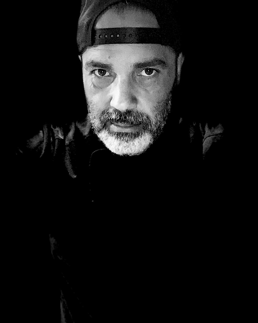 Ritratto in bianco e nero di Pierpaolo Grangilia, titolare e cuoco di Belgusto: berretto girato all&rsquo;indietro, barba brizzolata, in penombra su fondo nero
